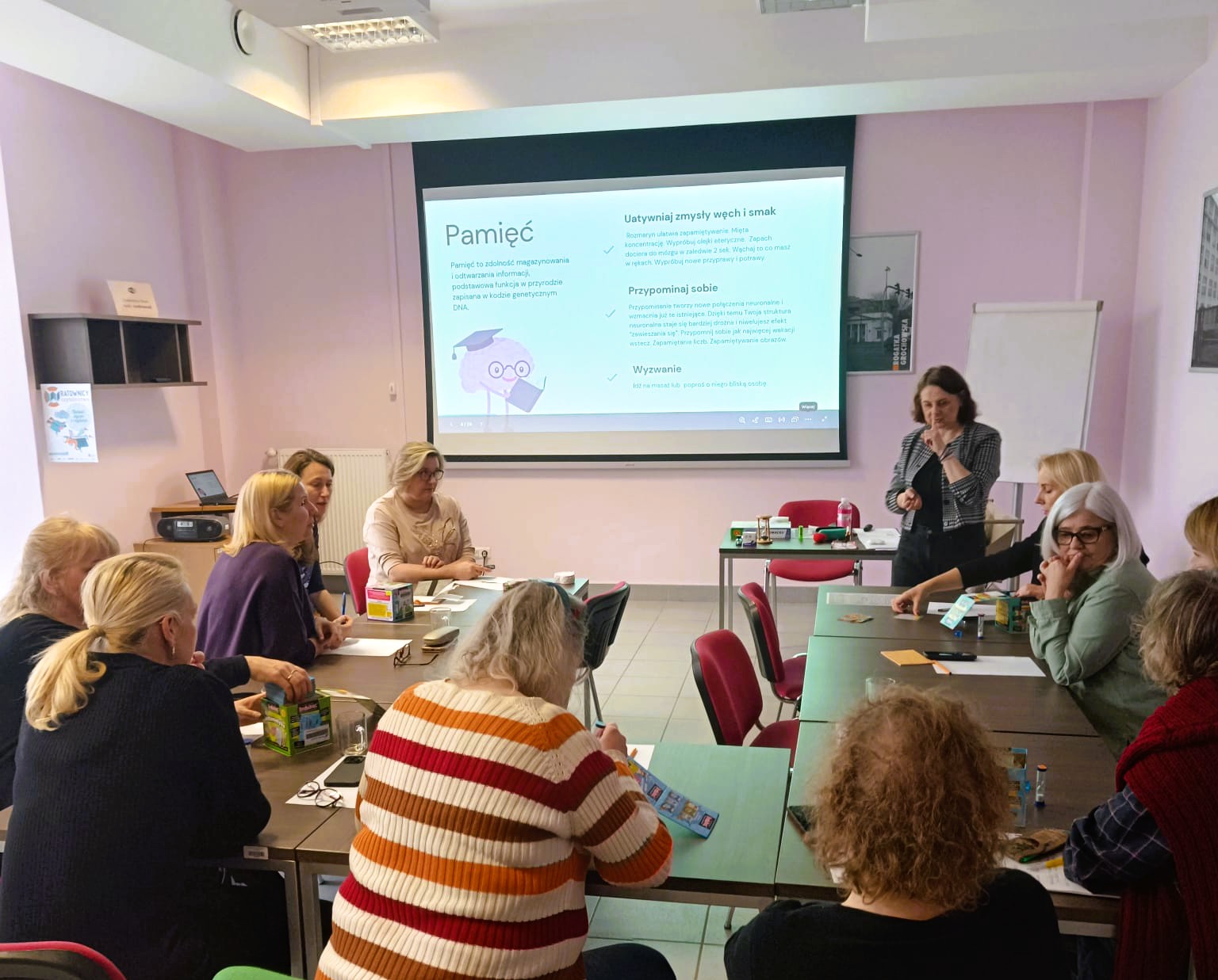 grupa ludzi siedzących przy stoliku wykonuje ćwiczenia pod kierunkiem osoby prowadzącej zajęcia ma tle wyświetlanej prezentacji