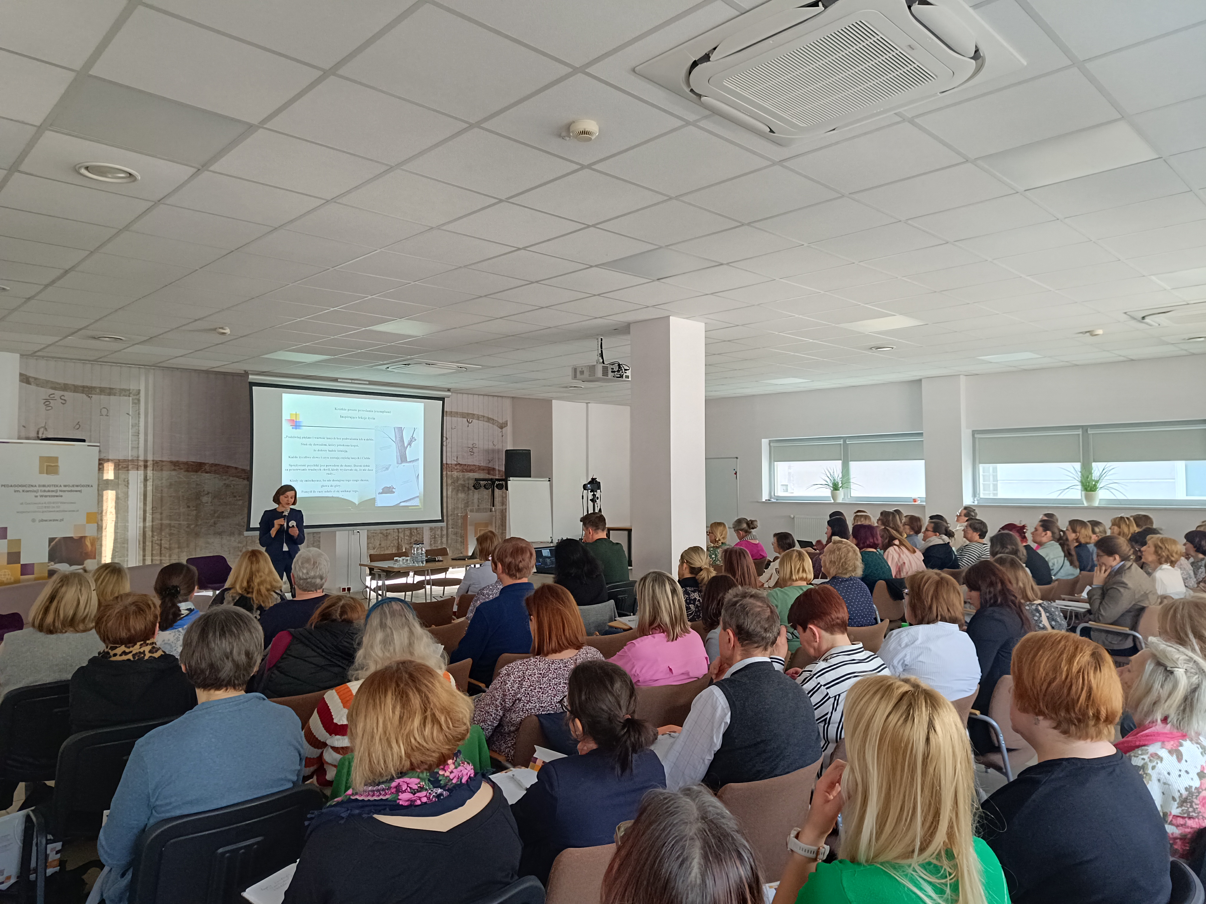 sala pełna ludzi, kobieta stoi na środku przed wszystkimi w tle prezentacja