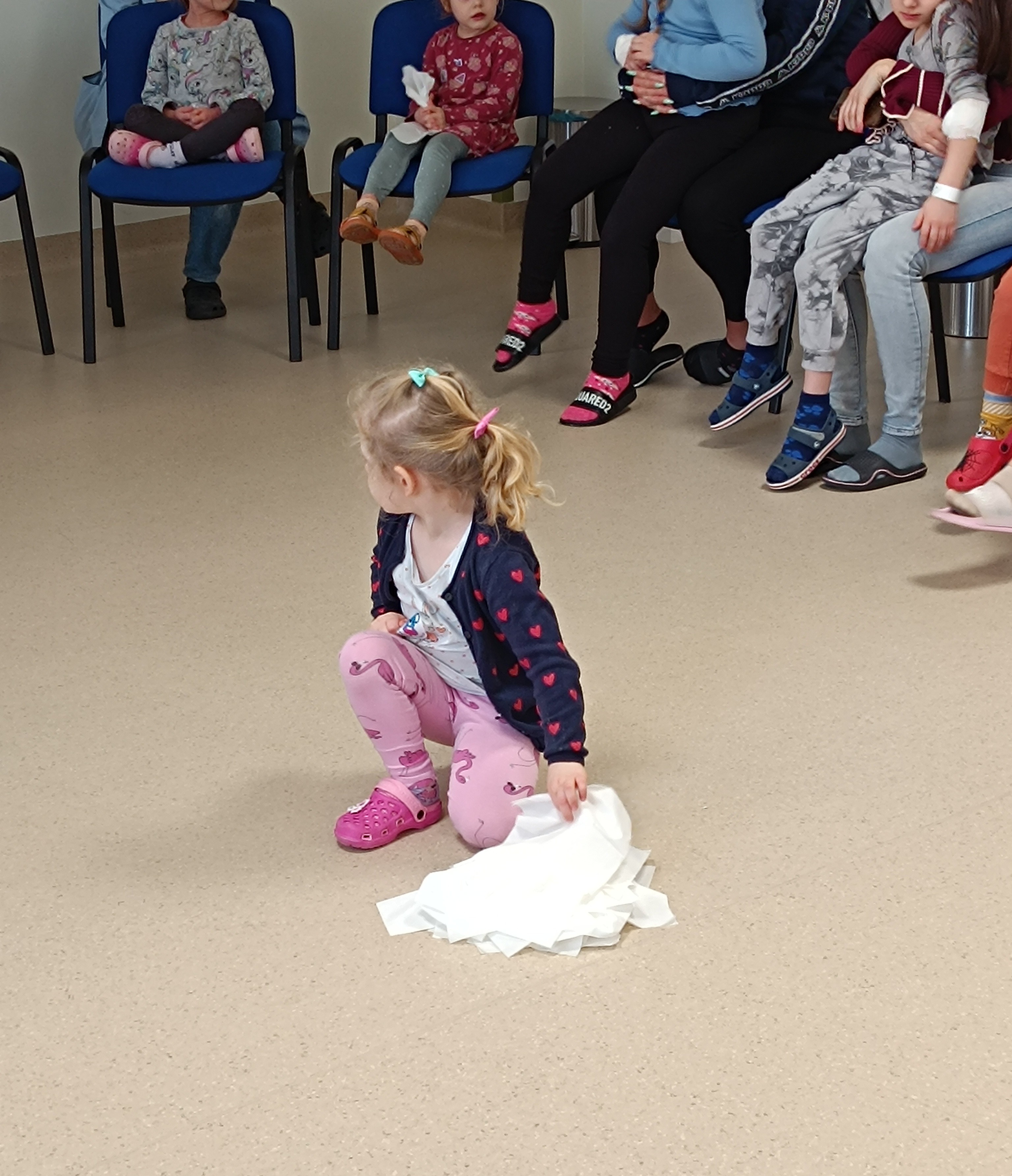 dziewczyka klęcząć na kolanie trzyma w prawej ręce biały materiał leżący częściowo na podłodze a otaczają ją siedzące na krzesłach osoby w różnym wieku, głównie dzieci 