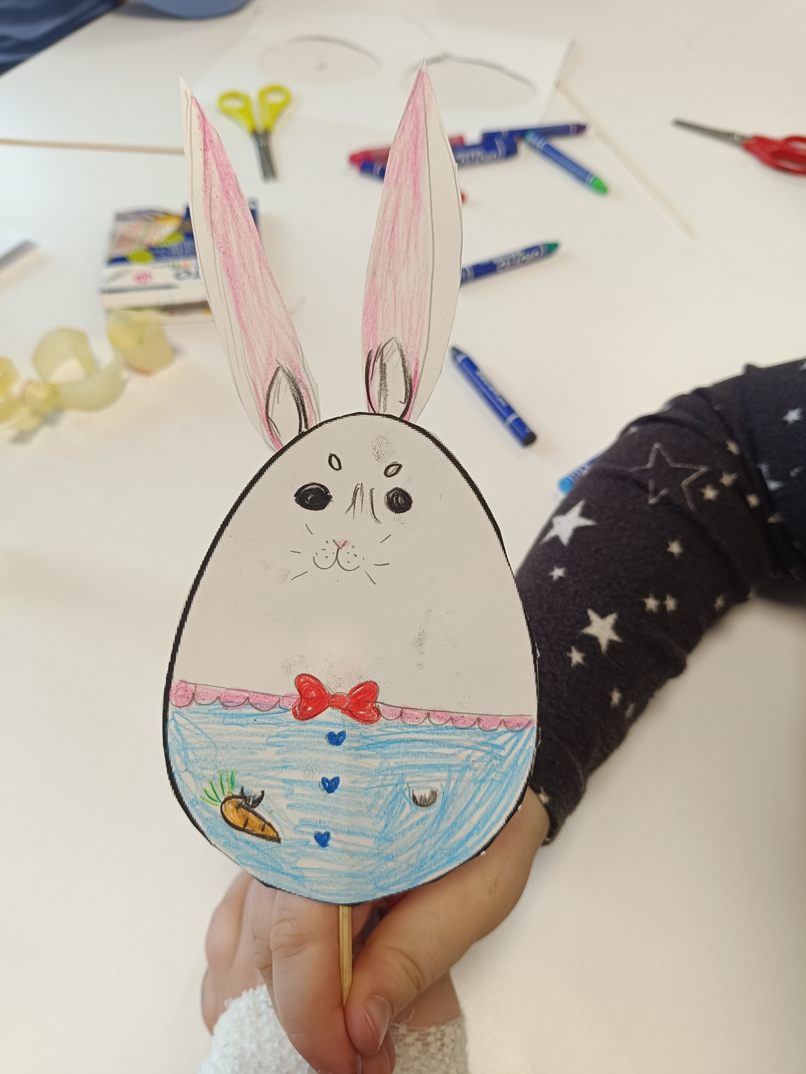 Zajączek w kształcie jajka narysowany przez dziecko, wycięty i przyklejony do patyczka trzymany ręką twórcy a w tle narzędzia do rysowania