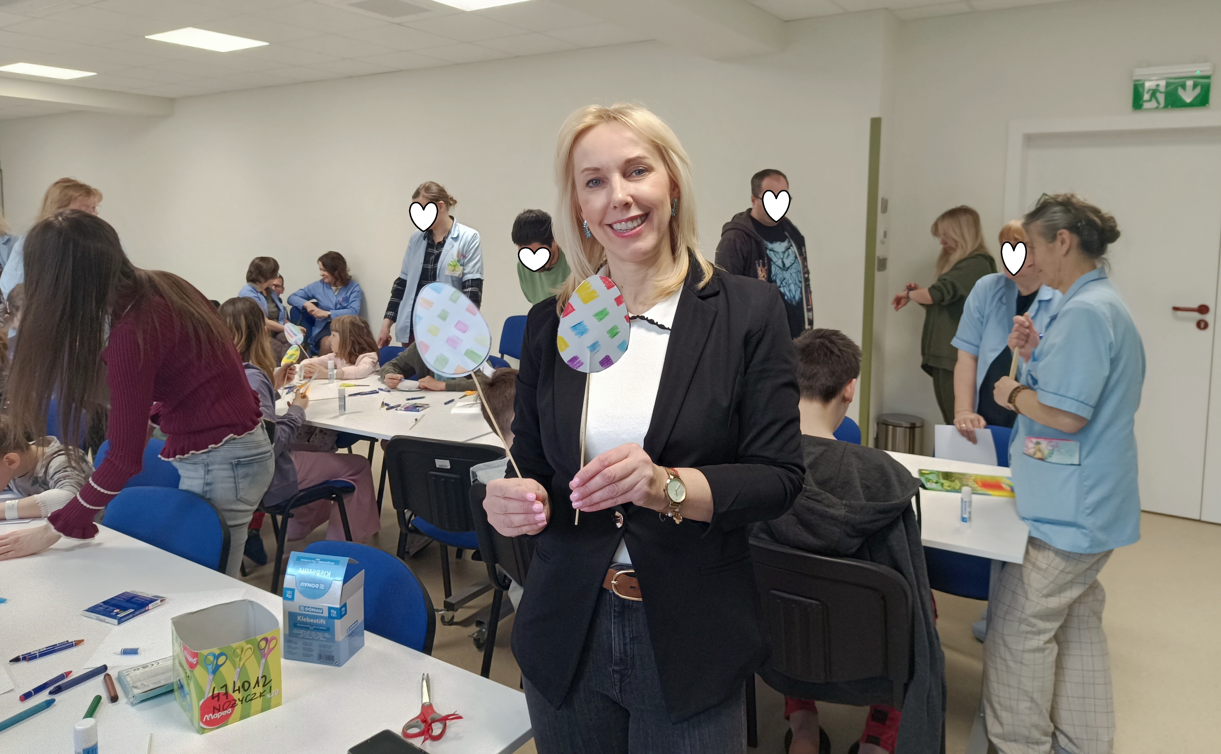 Prowadząca zajęcia pojazuje dwie kolorowe pisanki na patyczkach stworzone przez uczestników, których widać w tle przy stolikach wraz z dorosłymi opiekunami