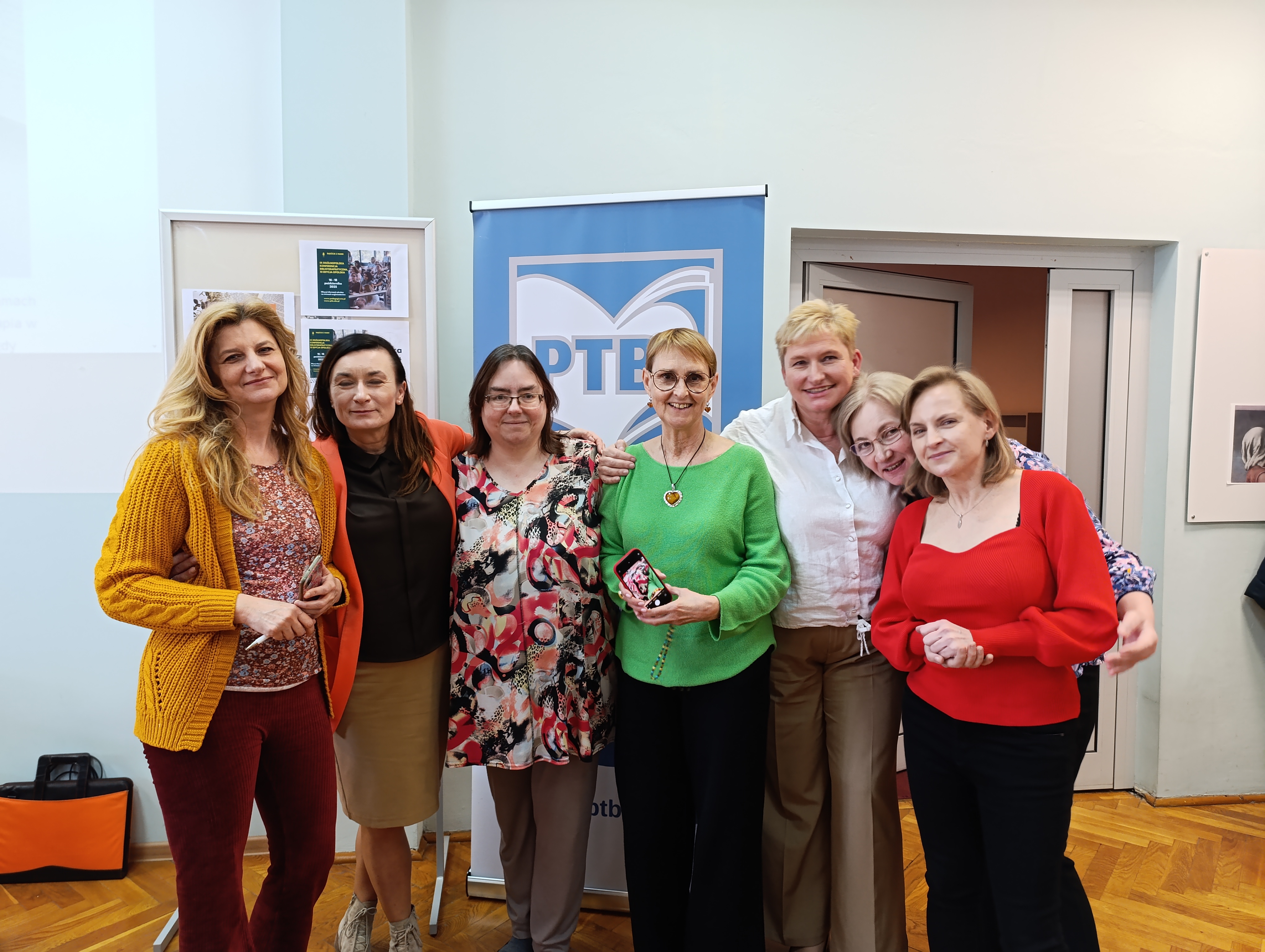 grupa kolorowo ubranych kobiet pozuje do zdjęcia na tle rollupa Polskiego Towarzystwa Biblioterapeutycznego
